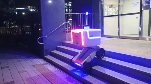 ALT Text: A delivery Robot going outside down a small flight of stairs on wheels with bright lights around the robot 