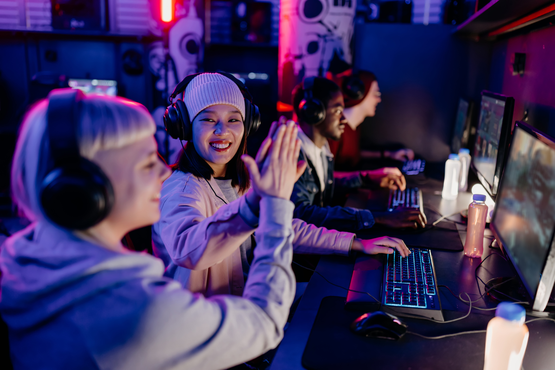 ALT Text :Four people wearing headsets are playing video games on computers in a dimly lit room. Two of them are high-fiving each other.