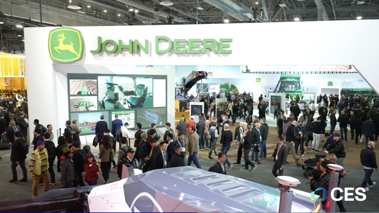 ALT Text: Conference full of people with a big arch with John Deere displayed on top 